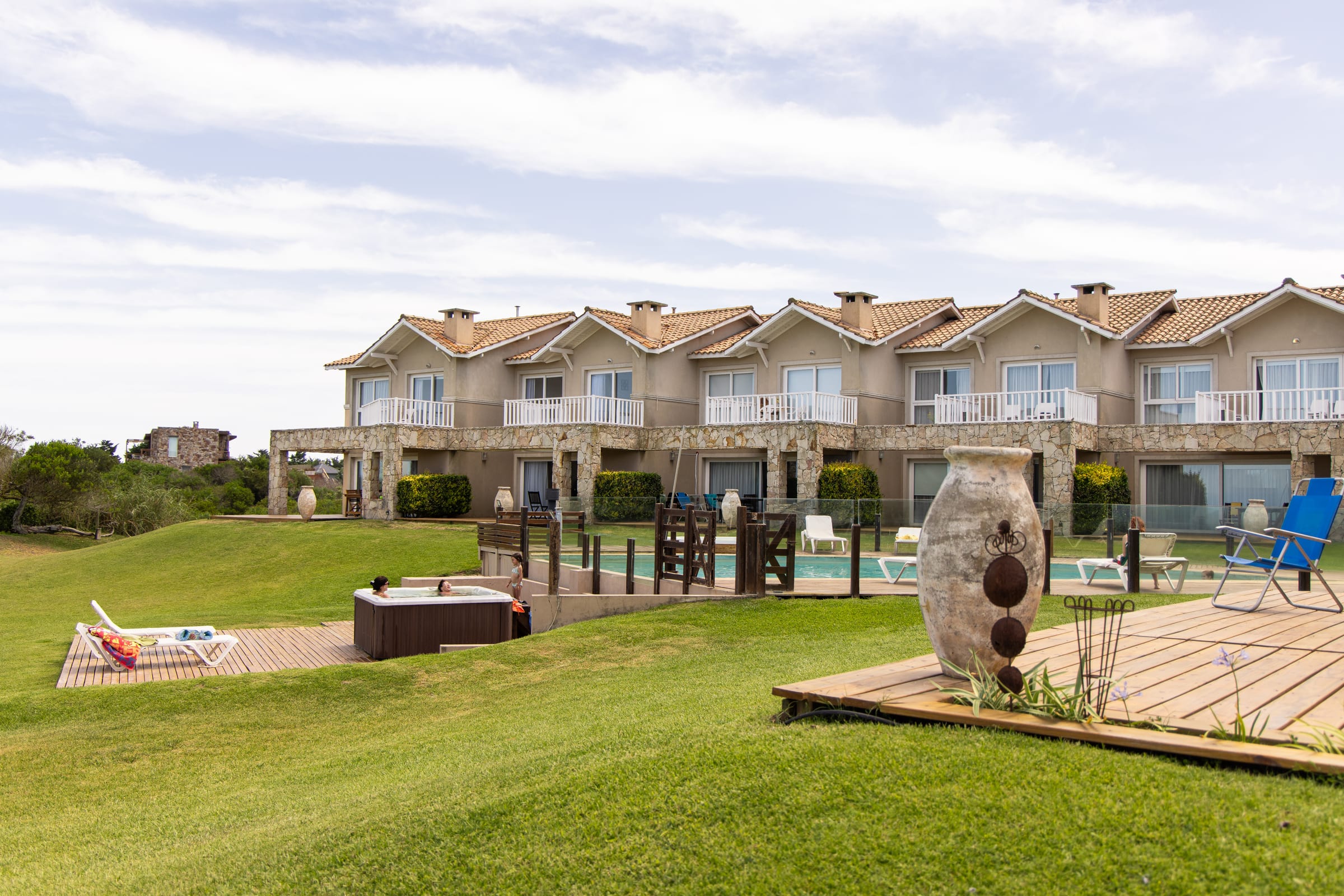 El deck de madera con el jacuzzi y las reposeras sobre el césped, las casas de piedra detrás.
